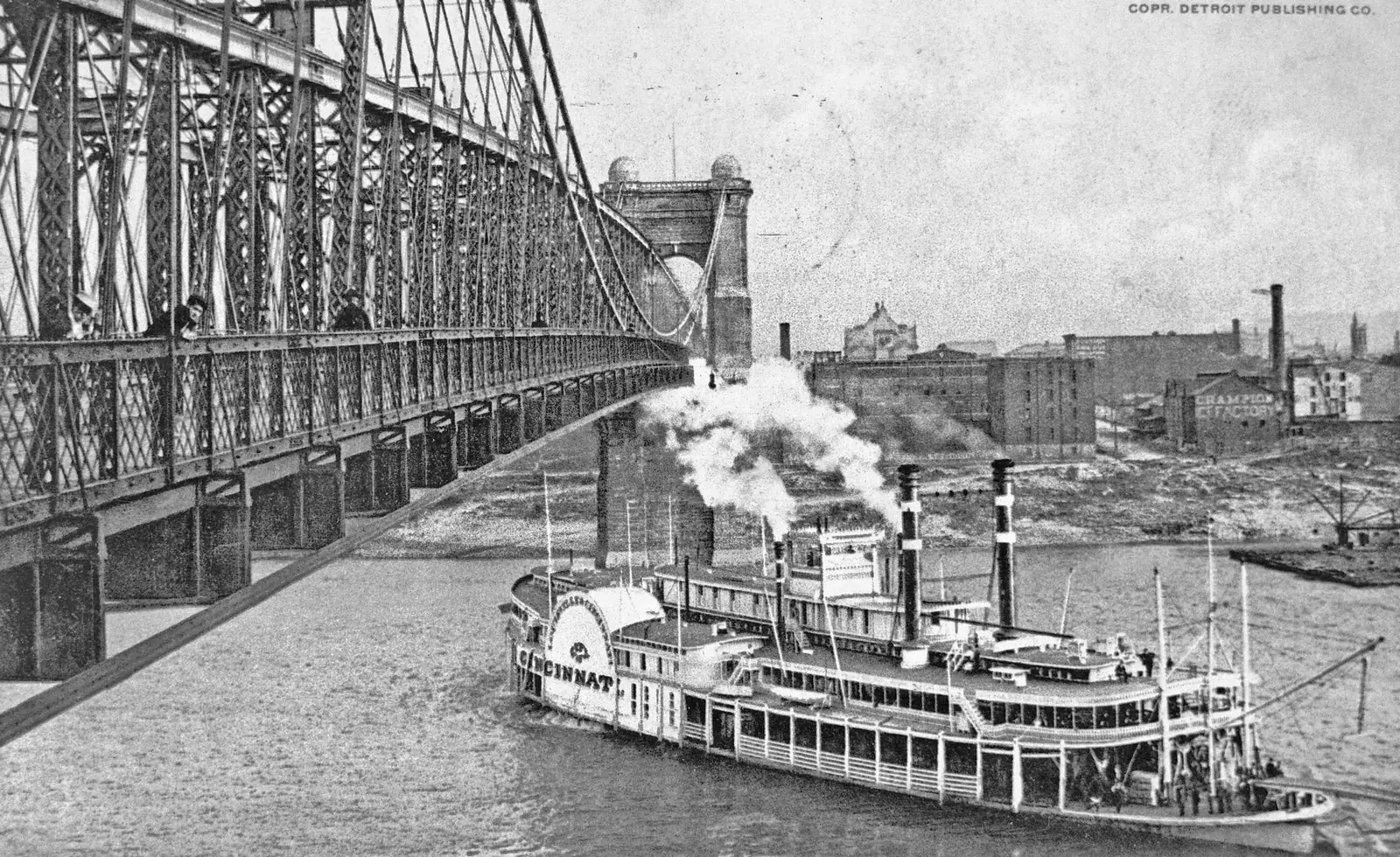 Sidewheel steamboat Cincinnati passing under the Roebling Suspension Bridge, riverfront factories behind, around 1900