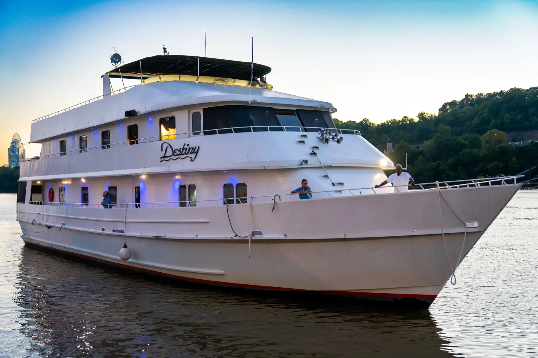 The Destiny Yacht on the Ohio River at dusk, her name on the hull and the Cincinnati skyline behind her